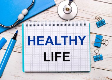 Hafif ahşap bir arka planda, steteskop, mavi bir not defteri, mavi ataç, mavi bir kalem ve Healtha Life yazılı bir kağıt. Tıbbi konsept