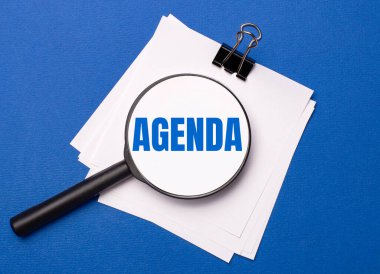 Mavi arka planda, siyah bir ataç altında beyaz çarşaflar ve üzerinde AGENDA metni olan bir büyüteç.