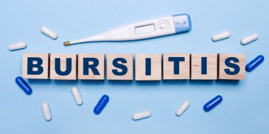 Açık mavi arka planda, elektronik termometre, beyaz ve mavi haplar ve üzerinde BURSITIS yazan tahta küpler. Tıbbi konsept