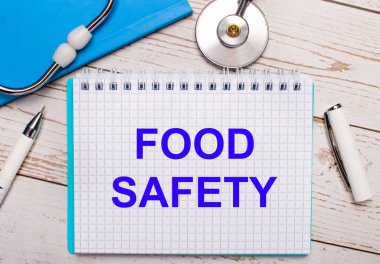 Hafif ahşap bir arka planda steteskop, mavi bir defter, beyaz bir kalem ve üzerinde FOOD Güvenlik yazıları olan bir kağıt var. Tıbbi konsept