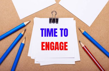 Açık kahverengi arka planda, mavi ve kırmızı kalemler ve Time to ENGAGE yazılı beyaz kağıt
