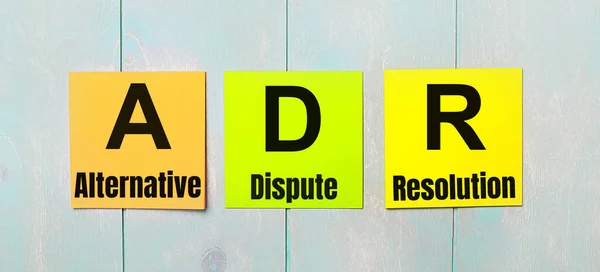 Three colored stickers with the text ADR Alternative Dispute Resolution a light blue wooden background