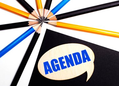 Çok renkli kalemlerin yanında siyah arkaplan üzerinde AGENDA yazan tahta kart.