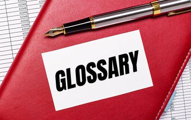 Masaüstünde raporlar, bordo bir defter, dolmakalem ve GLOSSARY yazılı beyaz bir kart var. İş konsepti