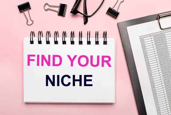 Pembe arka planda, raporlar, siyah ataçlar, gözlükler ve beyaz bir defter var.