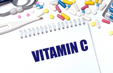 Hafif bir arka planda, çok renkli haplar, steteskop, elektronik termometre ve VITAMIN C yazılı bir defter. Tıbbi konsept..