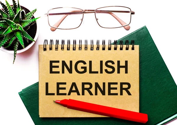 Hafif bir arkaplanda, altın çerçeveli bardaklar, saksıda bir çiçek, yeşil bir defter, kırmızı bir kalem ve üzerinde ENGLISH LEARNER yazan kahverengi bir defter. İş konsepti