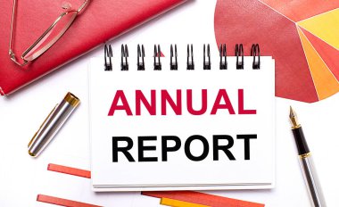 Masaüstünde ANNUAL REPORT yazılı beyaz bir defter, bir kalem, bordo ve kırmızı masalar ve altın çerçeveli gözlük var. İş konsepti.