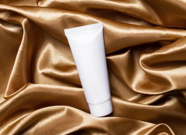 Kıvrımlı altın bir kumaşın arka planında beyaz bir krema tüpü var. Metni sokacak bir yer var. Güzellik ve sağlık kavramı