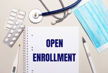 Açık gri bir arka planda, açık mavi tek kullanımlık yüz maskesi, stetoskop, elektronik termometre, haplar, bir kalem ve üzerinde ENROLMENT yazan bir defter. Tıbbi konsept