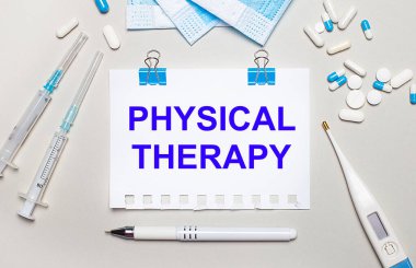 Açık gri arka planda, mavi tıbbi maskeler, şırıngalar, bir elektronik termometre, haplar, bir kalem ve üzerinde PHYSICAL THERAPY yazan bir defter. Tıbbi konsept