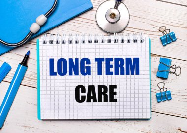 Hafif ahşap bir arka planda, steteskop, mavi bir not defteri, mavi ataç, mavi bir kalem ve uzun vadeli bakım tıbbi konsepti yazılı bir kağıt.