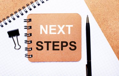 Tahta arka plan not defteri, siyah kalem, ataç ve NEXT STEPS yazılı kahverengi not defteri. İş konsepti