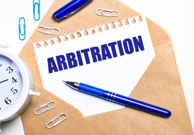 Hafif bir arkaplanda, bir el işi zarfı, bir çalar saat, ataçlar, mavi bir kalem ve ARBITRATION yazılı bir kağıt.