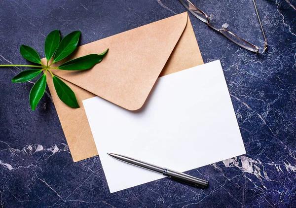 Koyu mermer bir arka planda, bir el işi zarfı, bir bitki yaprağı, bir kalem ve bir beyaz kağıt, içine yazı eklenecek bir yer. Şablon. Boşluğu kopyala