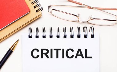 Hafif arkaplanda altın çerçeveli gözlükler, bir kalem, kahverengi ve kırmızı not defterleri ve CRITICAL yazılı beyaz bir defter. İş konsepti