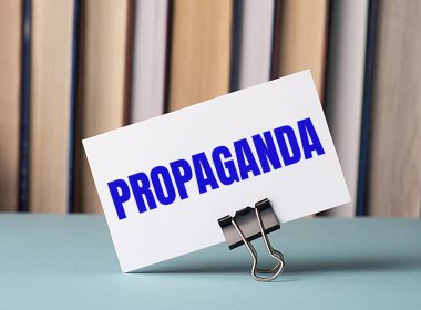 Üzerinde ProPAGANDA metni olan beyaz bir kart, kitapların arka planına karşı masada duran kağıtlar için bir şarjör üzerinde duruyor. Odaklanmayın