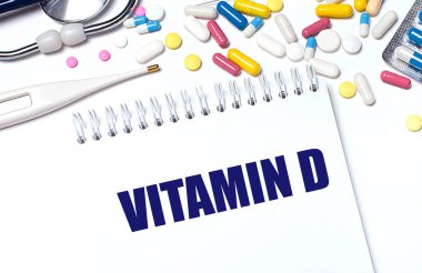 Hafif bir arka planda, çok renkli haplar, steteskop, elektronik termometre ve VITAMIN D yazılı bir defter. Tıbbi konsept..