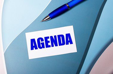 AGENDA yazılı beyaz kart ve mavi, mavi ve pembe arka planda mavi kalem.