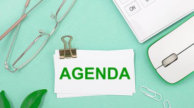 Açık yeşil arka planda beyaz bir hesap makinesi, bir bilgisayar faresi, bir bitkinin yeşil yaprakları, altın çerçeveli gözlük ve AGENDA yazılı beyaz bir kart var. İş konsepti