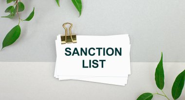 Gri arkaplanda bitkinin yeşil yaprakları ve altın bir şarjörün altında SANCTION LİST yazılı beyaz bir kart var. Minimalist.