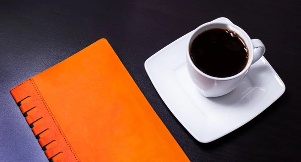 Μαύρο Φόντο Ένα Λευκό Φλιτζάνι Καφέ Και Ένα Πορτοκαλί Ημερολόγιο — Φωτογραφία Αρχείου