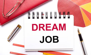 Masaüstünde DREAM JoB, kalem, bordo ve kırmızı masalar ve altın çerçeveli gözlüklerle dolu beyaz bir defter var. İş konsepti.