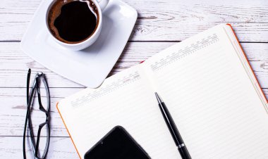 Açık ahşap bir masanın üzerinde açık bir defter, bir kalem, bir akıllı telefon, siyah çerçeveli bardaklar ve bir fincan kahve var. İş yeri kapanıyor.