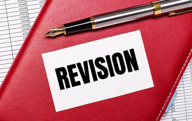 Masaüstünde raporlar, bordo bir defter, dolmakalem ve içinde REVISION yazan beyaz bir kart var. İş konsepti