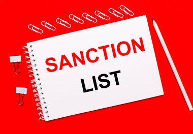 Parlak kırmızı bir arkaplanda, beyaz bir kalem, beyaz ataçlar ve SANCTION LİST yazılı beyaz bir defter.