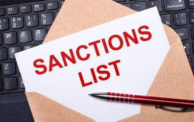 Üzerinde SANCTIONS LIST yazan beyaz kart çalışma masasındaki bir zarf içinde modern bir dizüstü bilgisayar klavyesi ve bordo kalemle birlikte. İş yerinin düz düzeni.