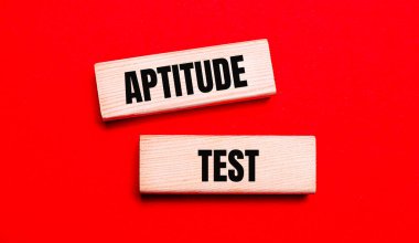 Parlak kırmızı arkaplanda, iki açık tahta blok var. Metin APTITUDE TEST