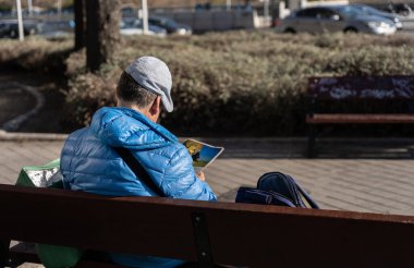 Adam parkta oturmuş dergi okuyor.