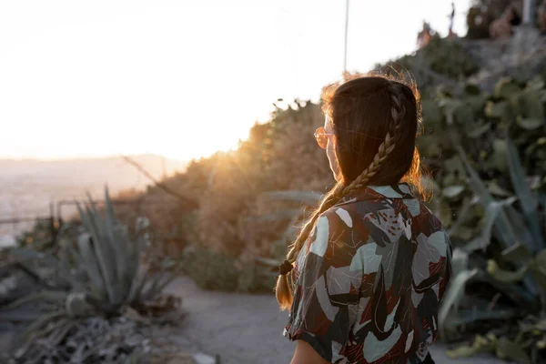 Unrecognizable young woman with ponytail and sunglasses is watching the sunset wearing a camouflage shirt