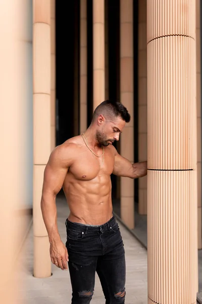 Üstsüz formasız bir adamın dikey fotoğrafı. Güçlü bir adam antrenmandan sonra kollarını bir sütuna dayayıp kaslarını esnetiyor.