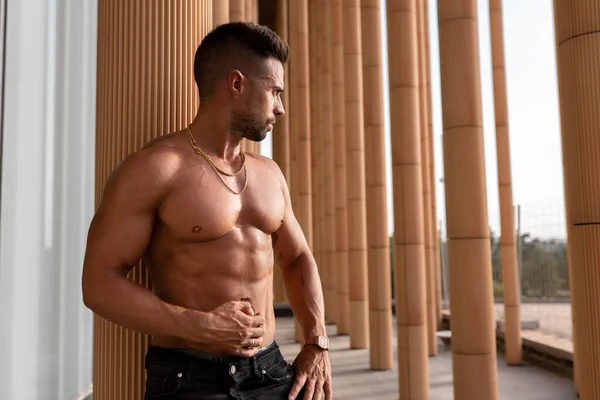 Üstsüz fitness. Güçlü bir adam dışarıda antrenman yaptıktan sonra karın kaslarına dokunarak bir sütuna yaslanıyor.