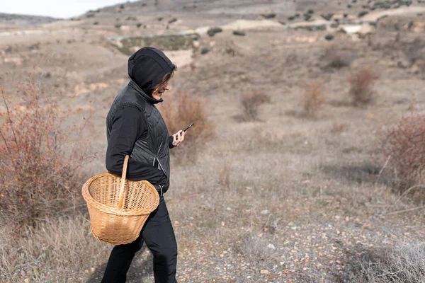 Mujer con una sudadera con capucha está mirando su teléfono mientras sostiene una cesta para recoger setas. — Foto de Stock