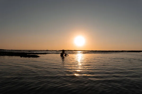 Yaz günbatımında denizde banyo yapan genç bir adamın silüeti.