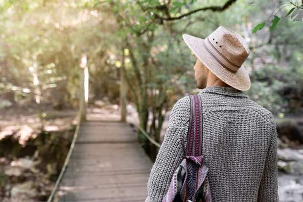 Şapka takan ve çanta taşıyan genç adam ormanın ortasında tahta bir köprüyü geçmek üzere. Doğada eğlenme kavramı