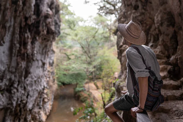 A young man wearing a hat is sitting on a high handrail and looking to a river flowing under him. Concept of adventure alone