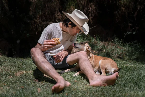 Şapka takan Meksikalı genç adam taco yiyor ve doğadaki çimlerin üzerinde köpeğiyle oynuyor. Boş zaman kavramı.