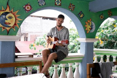 A young man is playing Spanish guitar outdoors sitting on the handrail of a balcony in Mexico. Concept of music artist traveler