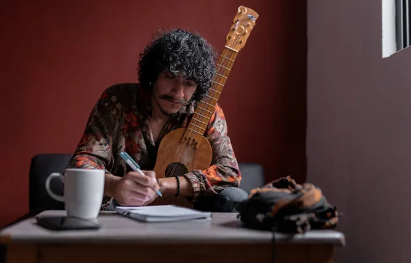 An young Mexican musician is writing a song in his notebook while holding his jarana guitar. Concept of practicing music.
