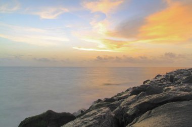 Sahilde gün batımında ön planda kayalarla çekilmiş uzun pozlu bir fotoğraf.