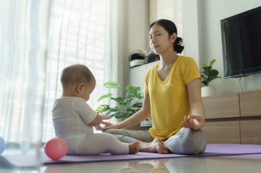 Asyalı genç anne, yoga yaparken yoga minderinde bebekle birlikte evdeki oturma odasında..