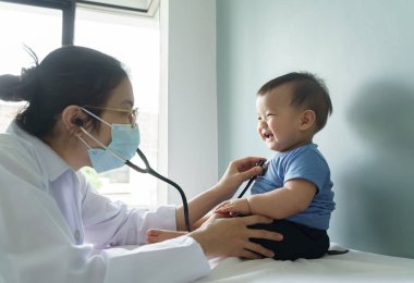 Yüz maskesi takan Asyalı kadın doktor hastanenin tıbbi odasında steteskoplu küçük tatlı çocuğu muayene ediyor..