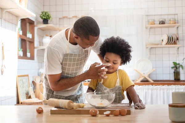 Happy smiling African American Father playing touching his little boy nose with flour while doing bakery at home, Black family having fun cooking baking cake or cookie in the kitchen together
