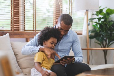Afro-Amerikalı baba ve küçük oğlu evdeki oturma odasında dijital tablet kullanıyorlardı. İnternet teknolojisiyle mutlu siyah aile.