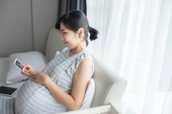 魅力的なアジアの若いです妊娠中の女性を保持し 見て超音波写真に座ってベッド — ストック写真
