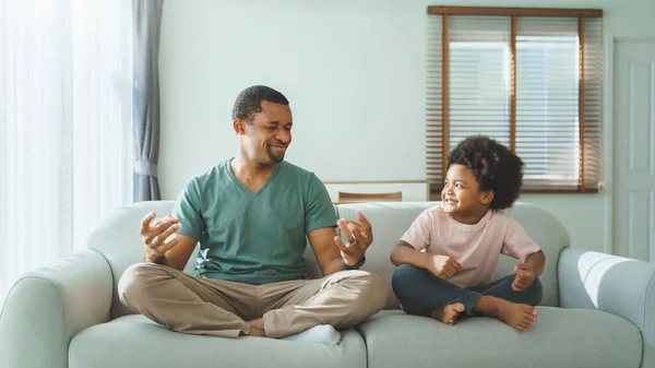 美国黑人父亲和快乐的小男孩坐在沙发上 一起在家里做瑜伽冥想 — 图库照片